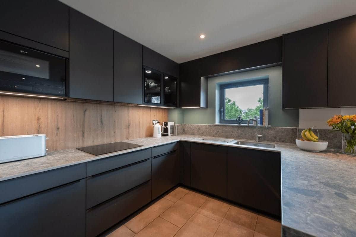 Modern dark German kitchen with handleless cabinetry and wood-effect splashback by Ebstone Kitchens